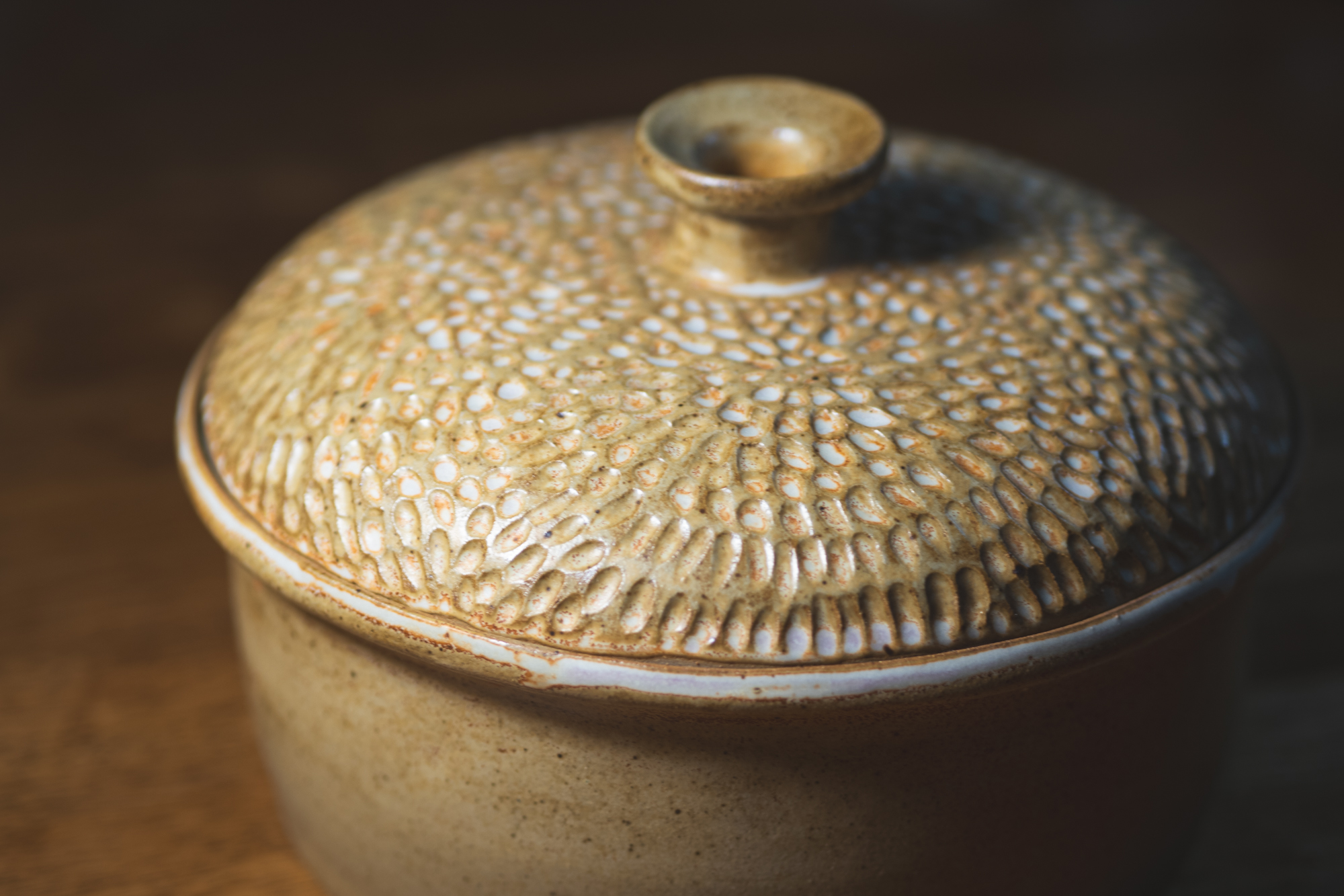 A carved stoneware casserole with a domed, hand-textured lid, glazed in honey ochre and resting on a walnut table in warm light.