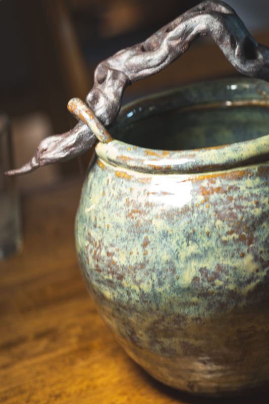 A mottled green vessel with a hand-sculpted branch form arching over its rim.
