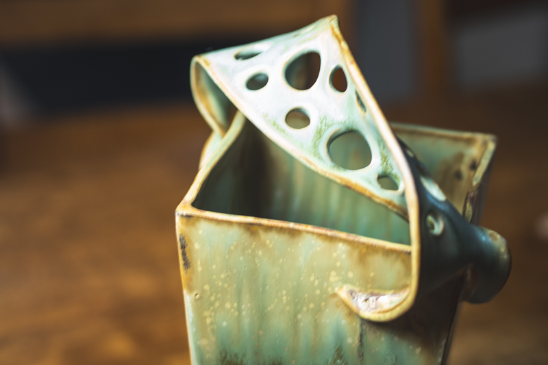 A square slab-built vase in sage green with a pierced, folded lid rising from one corner.