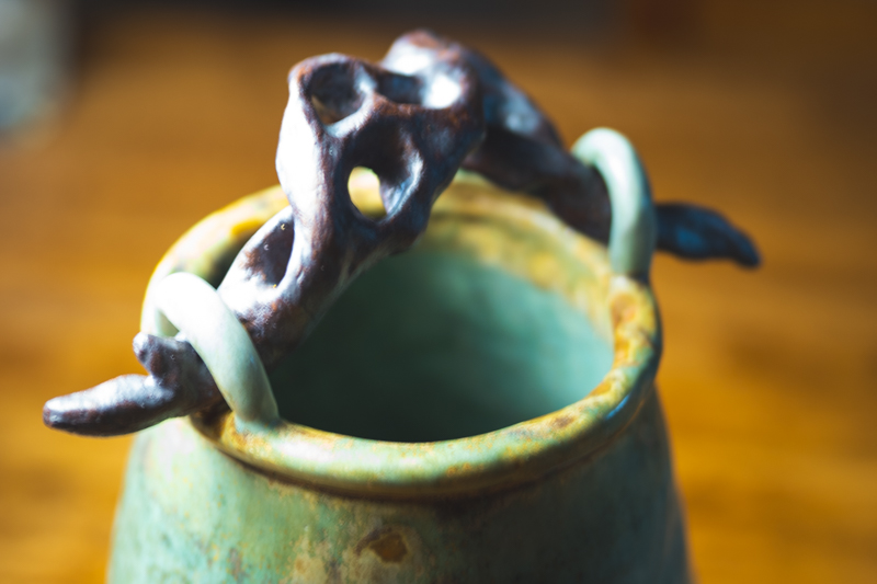Close detail of a sculpted root-like handle in dark glaze over a green vessel rim.