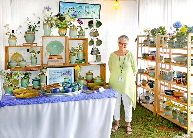 Elaine Agnew standing beside her booth at an outdoor show, surrounded by shelves of her green-glazed pottery and fresh flowers.