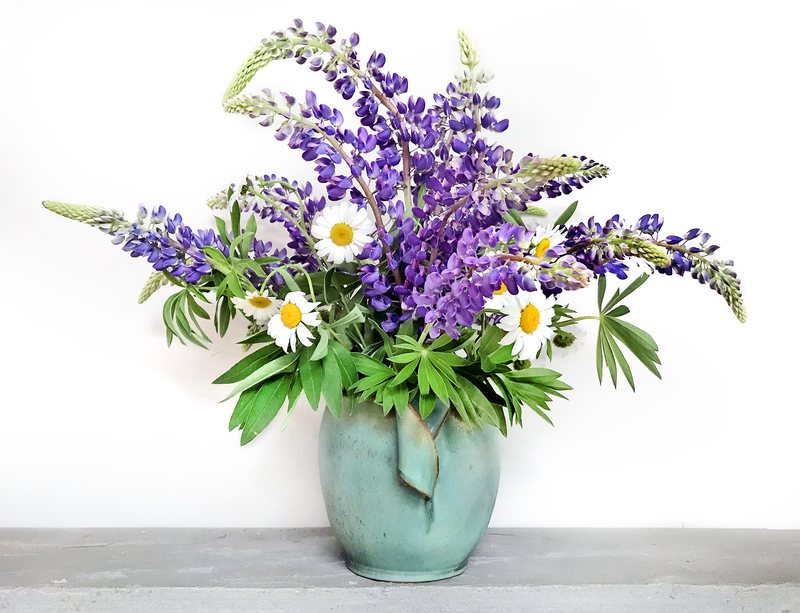 A small verdigris vase holding a wide arrangement of purple lupine and white daisies.