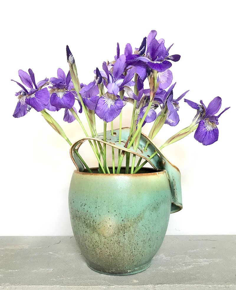 A rounded vase with a folded spout and strap handle, glazed in verdigris, holding purple iris.